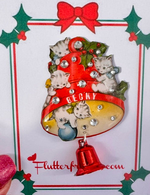 Christmas-themed pin or magnet with kittens and a red bell on a decorative card.