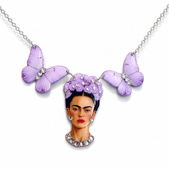 A silver necklace featuring a portrait of Frida Kahlo with purple flowers in her hair, two lavender butterfly charms, and rhinestone accents, shown on a white background.